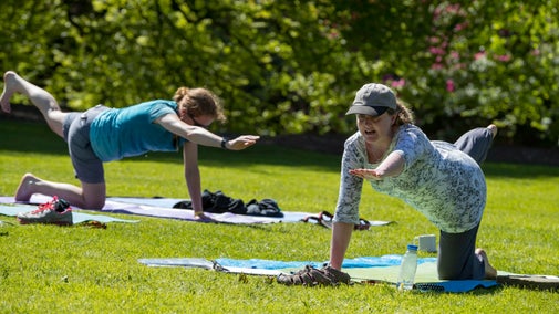 Two women are doing yoga positions, kneeling with one hand and the opposite foot in the air. They are on a green lawn surrounded by foliage.